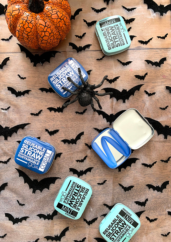 Gosili tins with straws lay on a brown background with bats, spiders, and a pumpkin. One of the tins is open and shows a blue reusable silicone straw. 