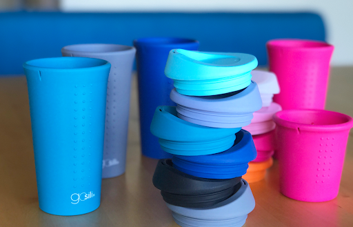 Reusable silicone cups in different vibrant colors standing next to each other. There's a stacked up tower of sustainable silicone tops in front of the cups.