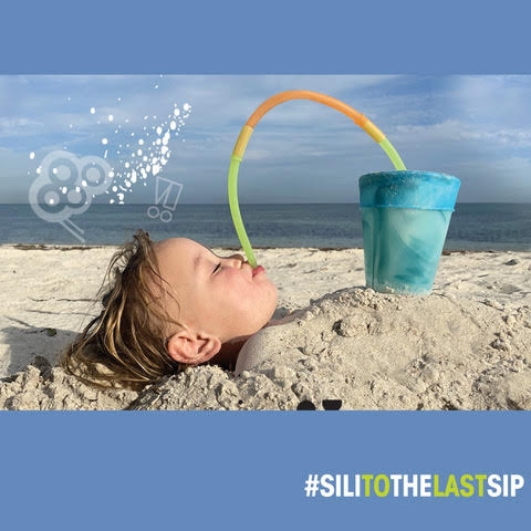 Kid buried in sand at the beach using a two connected silicone straws to drink from his silicone cup