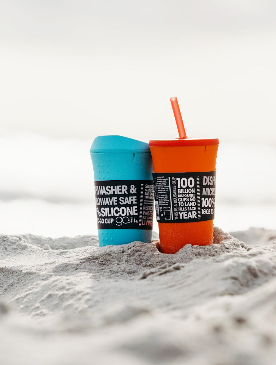 A blue reusable silicone cup with a sippy top and an orange silicone cup with an orange silicone straw sitting on the sand on a sunny day.